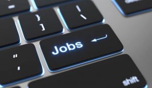 Um teclado de computador é mostrado em close, destacando uma tecla preta iluminada em azul com a palavra “Jobs” escrita no centro. A tecla possui o ícone de uma seta curva, semelhante ao de “Enter”, sugerindo uma função de acesso rápido relacionada a empregos. As outras teclas ao redor aparecem levemente desfocadas. (vagas)