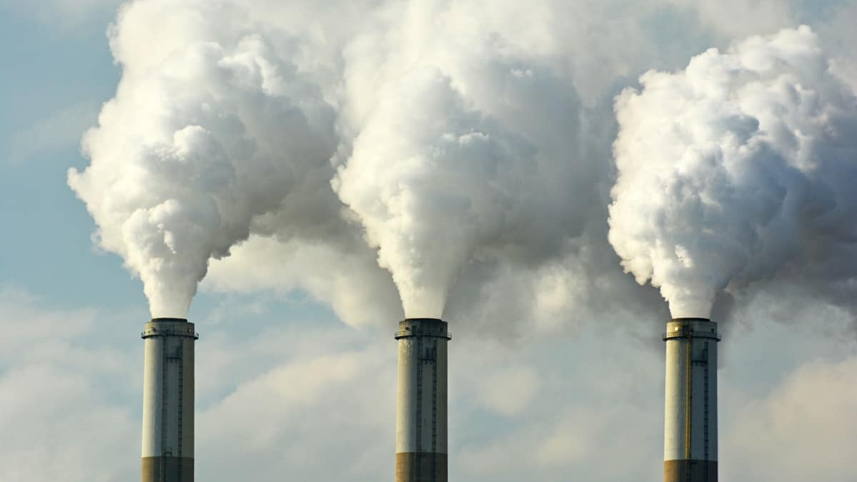 A imagem mostra três chaminés industriais emitindo grandes quantidades de fumaça branca ou vapor para o céu. As nuvens densas de emissões se espalham, contrastando com o fundo de um céu parcialmente nublado. A cena sugere poluição do ar e impactos ambientais relacionados à atividade industrial (startups, chaminés da indústria emitindo gás carbônico e poluição)