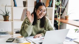 Jovem mulher sorridente com cabelos longos e óculos, usando um moletom verde, participando de uma videochamada em um notebook. Ela está sentada em uma mesa branca, acenando para a tela, com uma xícara de café, celular, caderno aberto e caneta ao seu lado. Ao fundo, há uma estante branca com livros e plantas decorativas, além de uma janela que deixa entrar luz natural (videochamada trabalho híbrido trabalho remoto)