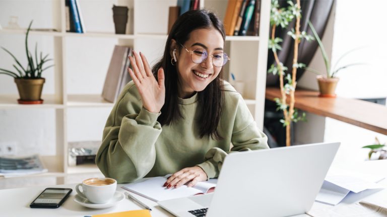 Jovem mulher sorridente com cabelos longos e óculos, usando um moletom verde, participando de uma videochamada em um notebook. Ela está sentada em uma mesa branca, acenando para a tela, com uma xícara de café, celular, caderno aberto e caneta ao seu lado. Ao fundo, há uma estante branca com livros e plantas decorativas, além de uma janela que deixa entrar luz natural (videochamada trabalho híbrido trabalho remoto)