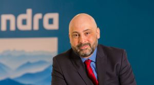 Marcelo Bernardino, CEO da Minsait no Brasil, está posicionado em frente a um fundo azul com o logotipo da empresa Indra. Ele veste um terno preto, camisa azul e gravata vermelha listrada, e exibe um sorriso sutil. A imagem transmite uma atmosfera profissional e corporativa