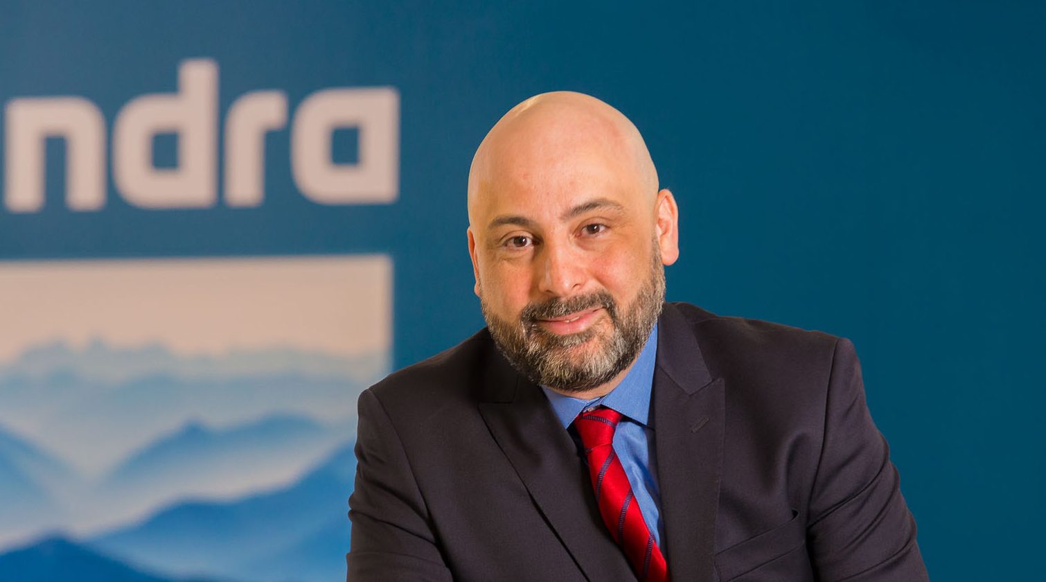 Marcelo Bernardino, CEO da Minsait no Brasil, está posicionado em frente a um fundo azul com o logotipo da empresa Indra. Ele veste um terno preto, camisa azul e gravata vermelha listrada, e exibe um sorriso sutil. A imagem transmite uma atmosfera profissional e corporativa