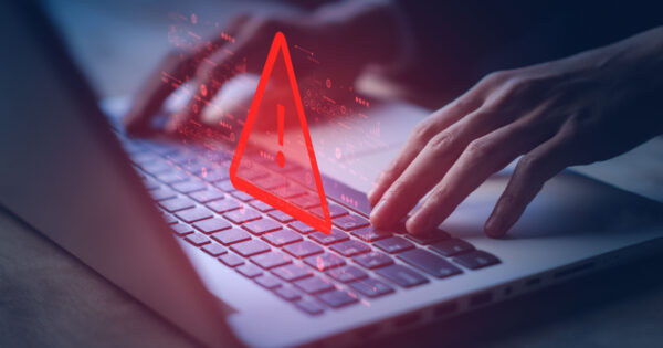 Imagem sombria de um laptop com destaque para um grande ícone vermelho de alerta no centro da tela, simbolizando uma ameaça digital. Há gráficos e elementos visuais em vermelho que remetem a dados e tecnologia, enquanto mãos digitam no teclado, sugerindo um possível ataque cibernético ou atividade maliciosa online (cibersegurança empresas OTRS, empresa, multiplan)