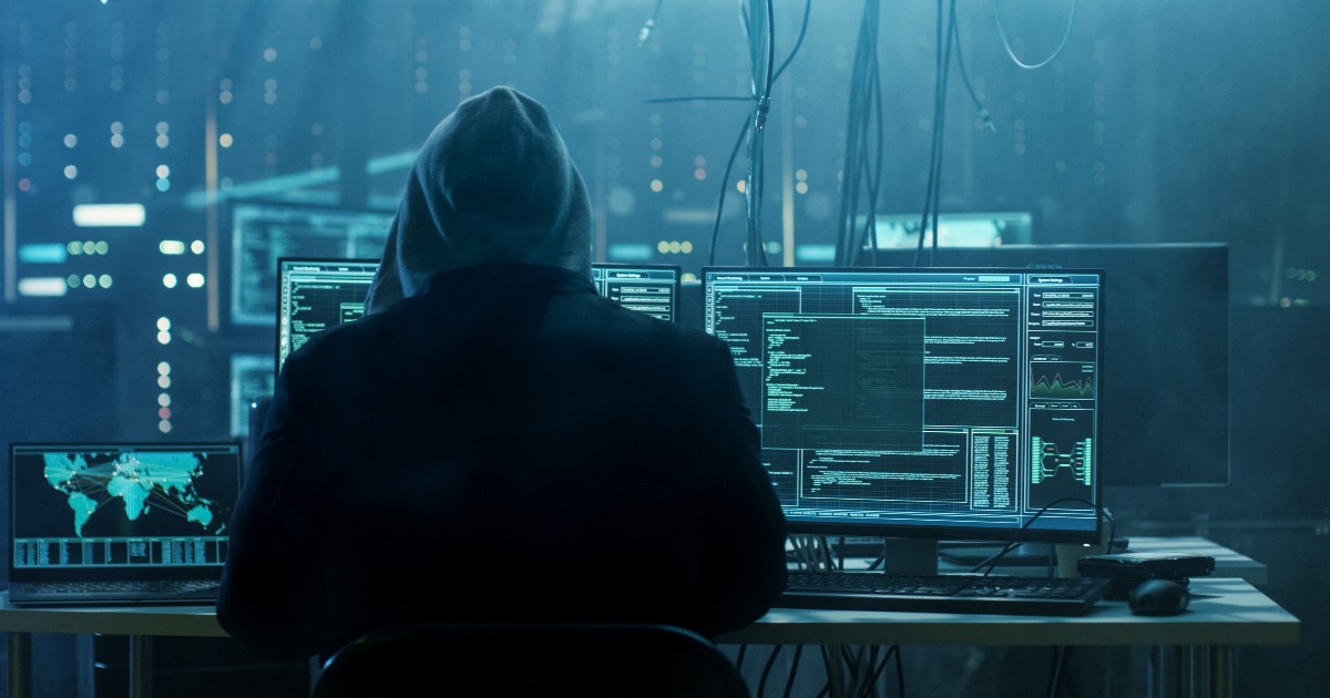 Indivíduo sentado em frente a múltiplas telas de computador em um ambiente escuro, com códigos e mapas globais exibidos nas telas, representando atividades de hacking ou segurança cibernética (cibercriminosos, hacker, cibercrimes, hp crimes cibernéticos, setor financeiro, finanças, bancos, dados, man-in-the-middle, europol)