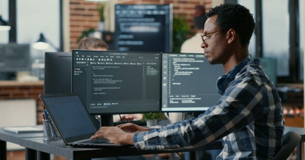 Homem negro usando óculos e camisa xadrez azul trabalha em um escritório moderno com três telas à sua frente, incluindo um notebook e dois monitores exibindo códigos de programação. Ele está concentrado no desenvolvimento de software, com fones de ouvido apoiados no pescoço. Ao fundo, há outros profissionais e um ambiente com tijolos aparentes e janelas amplas (desenvolvedores, OutSystems, desenvolvedores de software, Husky, cibersegurança, profissionais, software)