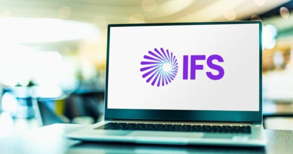 Imagem de um notebook sobre uma mesa, com a tela exibindo o logotipo da empresa IFS. O logotipo é composto por um símbolo em formato espiralado com linhas roxas e ao lado está a sigla “IFS” em letras maiúsculas, também na cor roxa. O fundo da imagem é desfocado, sugerindo um ambiente interno moderno e iluminado.