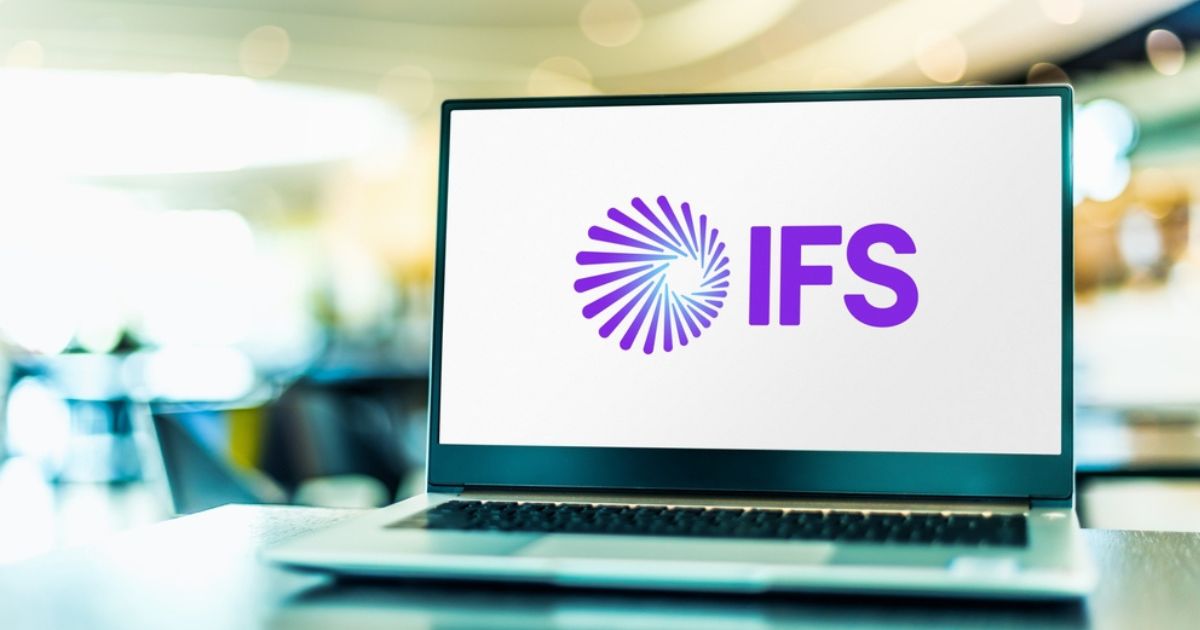 Imagem de um notebook sobre uma mesa, com a tela exibindo o logotipo da empresa IFS. O logotipo é composto por um símbolo em formato espiralado com linhas roxas e ao lado está a sigla “IFS” em letras maiúsculas, também na cor roxa. O fundo da imagem é desfocado, sugerindo um ambiente interno moderno e iluminado.
