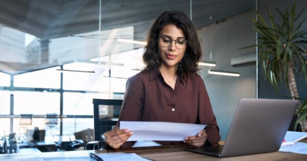 Mulher profissional em um escritório moderno, analisando documentos com atenção. Ao lado dela, há um laptop, representando um ambiente de trabalho focado e organizado, compras públicas, gestão documental, vagas, cursos, ransomware