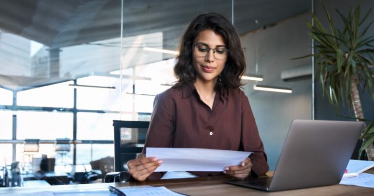 Mulher profissional em um escritório moderno, analisando documentos com atenção. Ao lado dela, há um laptop, representando um ambiente de trabalho focado e organizado, compras públicas, gestão documental, vagas, cursos, ransomware