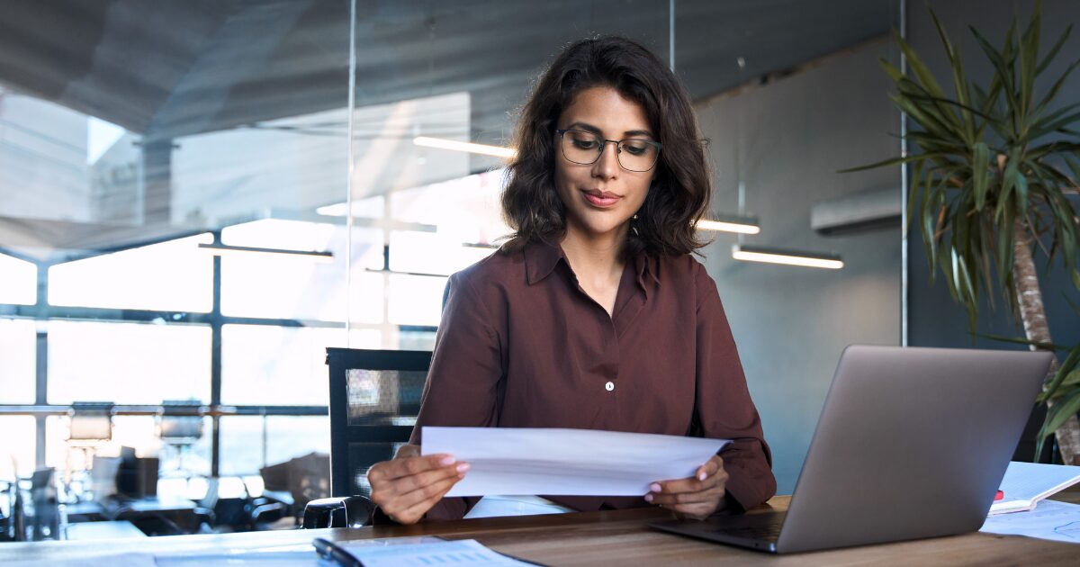 Mulher profissional em um escritório moderno, analisando documentos com atenção. Ao lado dela, há um laptop, representando um ambiente de trabalho focado e organizado, compras públicas, gestão documental, vagas, cursos, ransomware