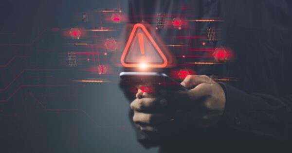 Pessoa segurando um smartphone com ícones e símbolos digitais em vermelho projetados no ar, incluindo um grande triângulo de alerta com um ponto de exclamação. A imagem simboliza um alerta de segurança cibernética ou vulnerabilidade em dispositivos móveis, com elementos gráficos digitais ao fundo (iot, ciberataque, vulnerabilidade, alerta, segurança, cibersegurança, dispositivo, device, golpe, ataques maliciosos, risco)