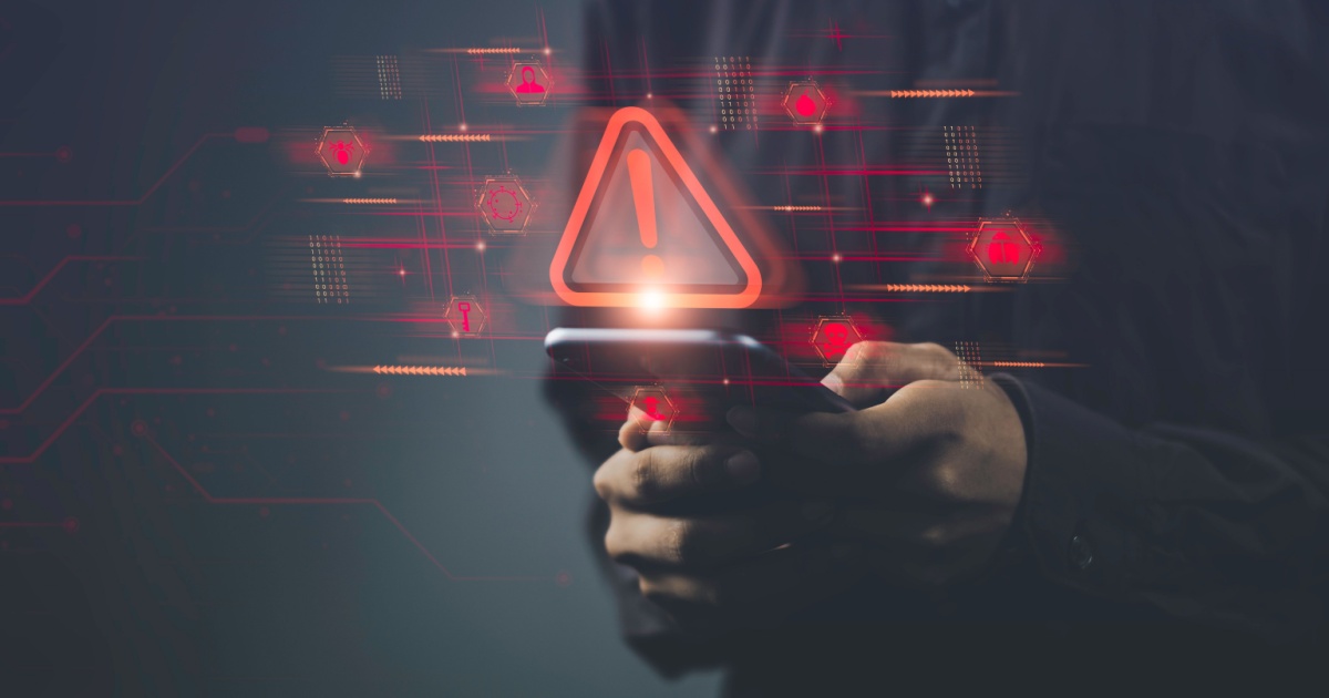 Pessoa segurando um smartphone com ícones e símbolos digitais em vermelho projetados no ar, incluindo um grande triângulo de alerta com um ponto de exclamação. A imagem simboliza um alerta de segurança cibernética ou vulnerabilidade em dispositivos móveis, com elementos gráficos digitais ao fundo (iot, ciberataque, vulnerabilidade, alerta, segurança, cibersegurança, dispositivo, device, golpe, ataques maliciosos, risco)