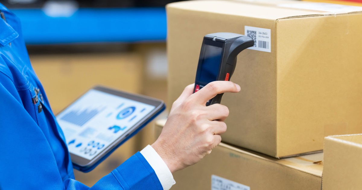 Trabalhador em um ambiente de logística utilizando um scanner de código de barras em uma caixa de papelão, enquanto segura um tablet com gráficos e dados exibidos na tela. Ele veste uniforme azul e está cercado por pacotes, sugerindo um processo de controle de estoque e rastreamento de produtos em um armazém (carbono, tecnologia)