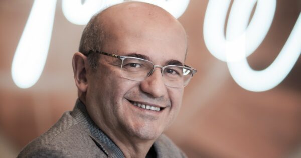 Cleber Morais, diretor-geral de vendas da AWS no Brasil, sorrindo para a câmera com um fundo desfocado em tons claros. Ele está usando óculos elegantes e um paletó cinza, transmitindo uma imagem de confiança e simpatia (IA generativa)