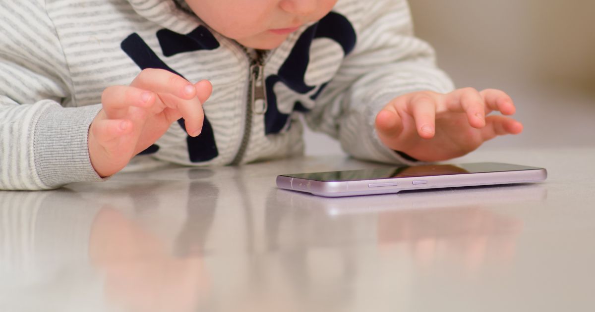 Esta imagem mostra uma criança pequena debruçada sobre uma superfície brilhante, interagindo com um smartphone. As suas mãos pequenas estão posicionadas perto do dispositivo, como se estivesse a tocar no ecrã ou a explorar o telemóvel. A criança veste um casaco cinzento com padrão listrado e um fecho de correr visível, e a cena sugere concentração ou curiosidade infantil. O foco está na interação entre a criança e o smartphone, representando um momento moderno e comum na infância atual. Kaspersky