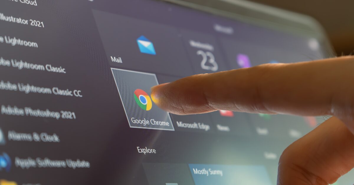 Dedo de uma pessoa tocando o ícone do navegador Google Chrome em uma tela de computador, que mostra vários ícones de aplicativos
