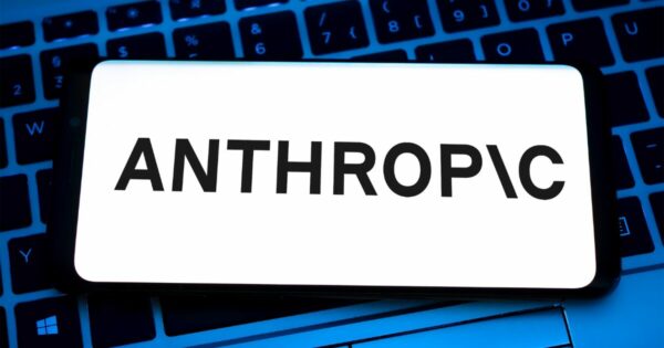 Imagem mostrando a tela de um smartphone com o logotipo da Anthropic em destaque, com letras pretas sobre fundo branco. Ao fundo, é visível um teclado de computador desfocado, em tons escuros e iluminação azulada, criando um contraste com a nitidez do logotipo; a empresa lançou ferramento IA para cibersegurança.