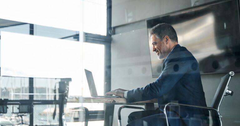 A imagem mostra um executivo de negócios sorrindo enquanto trabalha em seu laptop. Ele está sentado em uma cadeira de escritório em um ambiente moderno e luminoso, com grandes janelas ao fundo, oferecendo uma vista panorâmica. A cena transmite uma atmosfera de bem-estar, produtividade e liderança, destacando o executivo em um momento de foco e satisfação enquanto realiza suas tarefas no escritório (ceo, executivo, bem-estar, saúde mental, trabalho, liderança, diretor)