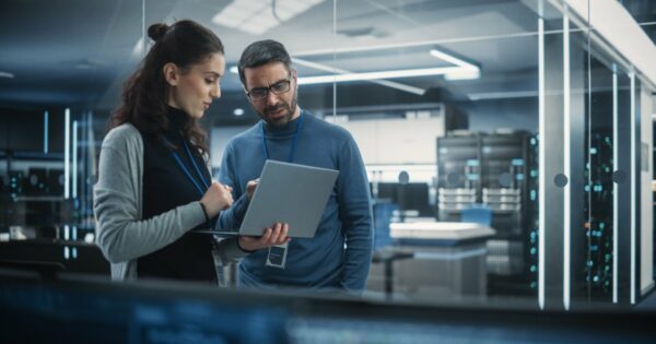 Dois profissionais de TI discutindo enquanto analisam informações em um laptop dentro de um data center moderno. O ambiente é iluminado por luzes azuis e cercado por servidores de alta tecnologia, com um cenário futurista e profissional, executivos, salários, empregos (Archimate, desenvolvedor)