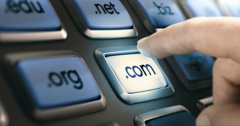 Close-up de um dedo pressionando um botão com o texto '.com', representando um domínio de topo (Top Level Domain - TLD). Outros botões ao redor exibem extensões como .org, .net e .biz, simbolizando registros de domínios na internet