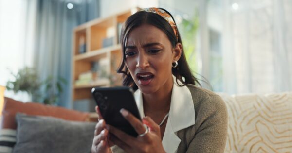 Mulher expressando surpresa ou preocupação enquanto olha para o celular, retratando o impacto de possíveis notícias ou informações inesperadas no ambiente doméstico, desinformação, fake news