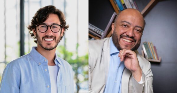 Uma pessoa com barba volumosa e escura veste um blazer azul‑marinho em um ambiente interno amplo e iluminado. Ao fundo, há diversas luzes retangulares no teto, criando um padrão linear, e janelas de vidro que revelam salas iluminadas. A cena sugere um espaço corporativo ou um centro de convenções durante um evento.