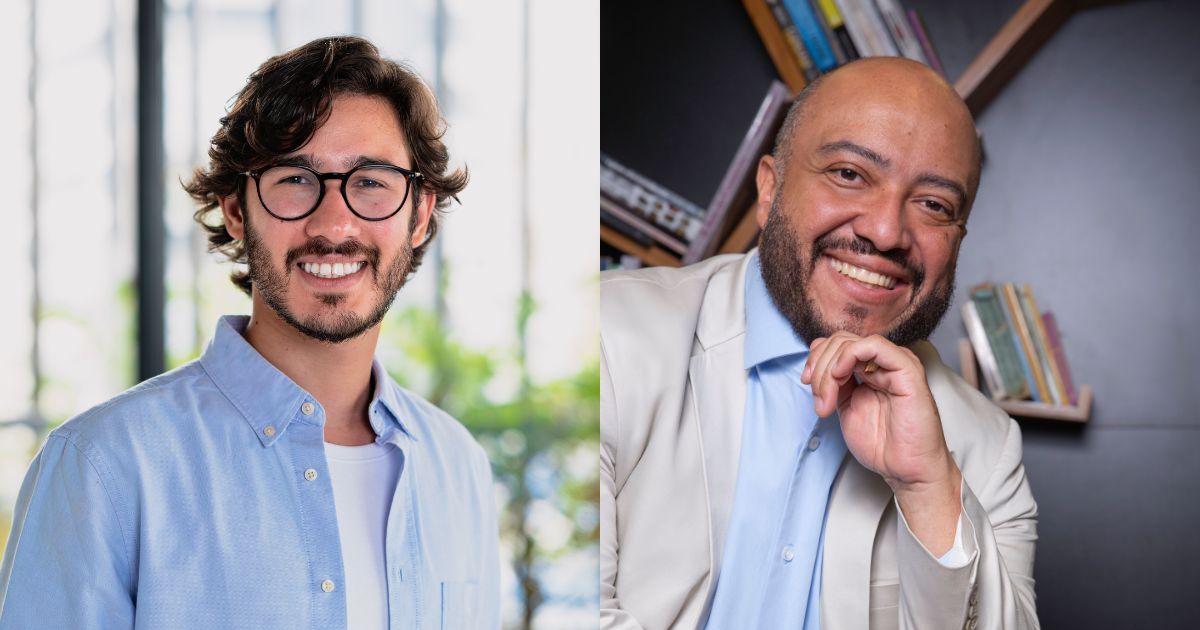 Uma pessoa com barba volumosa e escura veste um blazer azul‑marinho em um ambiente interno amplo e iluminado. Ao fundo, há diversas luzes retangulares no teto, criando um padrão linear, e janelas de vidro que revelam salas iluminadas. A cena sugere um espaço corporativo ou um centro de convenções durante um evento.