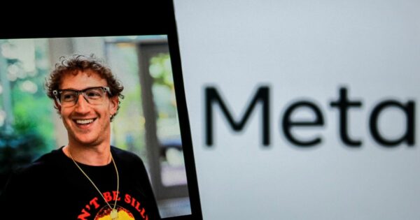 Uma fotografia mostra um homem sorridente com cabelo encaracolado e óculos, vestido de forma casual, com uma camisa preta estampada com um texto colorido. Ao lado dele, parcialmente visível, está o logótipo da empresa "Meta" sobre um fundo branco. A imagem combina o rosto do homem em destaque e o nome da empresa em plano de fundo.