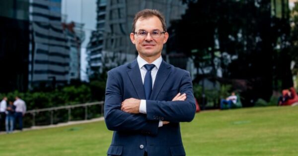 Imagem de Marcos Ferrari, presidente-executivo da Conexis, em um ambiente externo. Ele está usando um terno azul escuro com gravata, de braços cruzados, transmitindo uma postura confiante e profissional. Ao fundo, há um gramado verde, árvores e edifícios modernos, criando um cenário corporativo ao ar livre (Open Gateway)