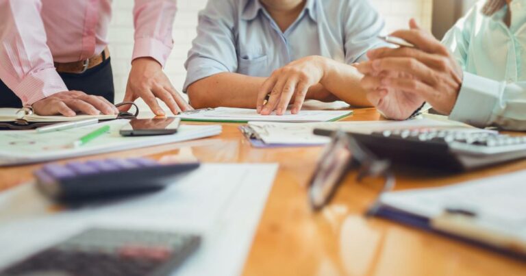 Imagem de um grupo de pessoas em uma mesa de reunião discutindo documentos e planilhas. A mesa está coberta com materiais de escritório, como calculadoras, cadernos, canetas e smartphones, simbolizando um ambiente de trabalho colaborativo e organização de negócios (ano novo, empresas)