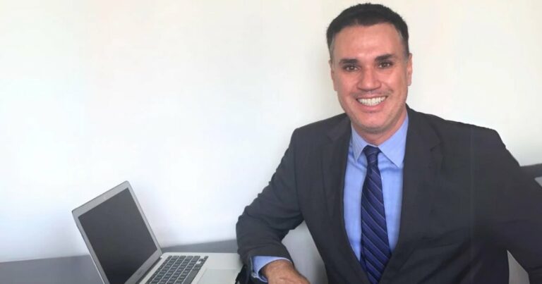 A imagem mostra Franklin Tomich, fundador da Accordia, sentado à mesa com um laptop ao lado. Ele veste um terno escuro, camisa azul e gravata listrada, transmitindo um visual profissional. Com um sorriso confiante, ele olha diretamente para a câmera. O fundo é simples, com uma parede branca, destacando o ambiente corporativo e minimalista da foto (Franklin Tomich, Accordia, risco, planejamento)
