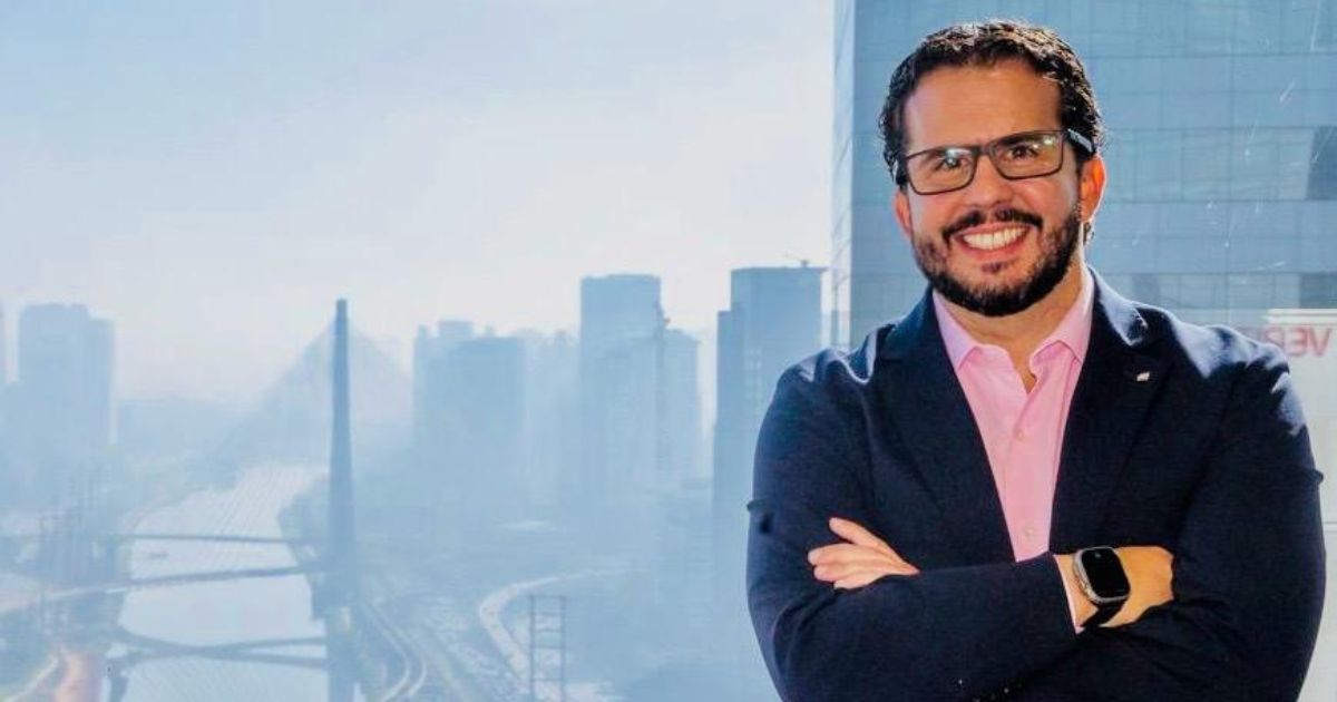 Homem sorridente com óculos e blazer azul posando em frente a uma janela com vista para a cidade de São Paulo, destacando a Ponte Estaiada e prédios ao fundo. Gustavo Leite, VP América Latina e Caribe da Cohesity. Foto: Divulgação