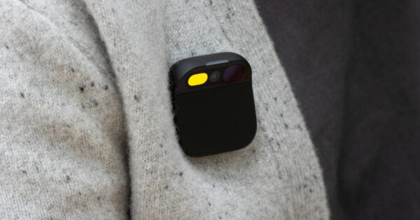 A imagem mostra um Humane Ai Pin, um dispositivo tecnológico compacto e vestível, fixado na roupa de uma pessoa. O aparelho tem um design quadrado com cantos arredondados e uma superfície preta fosca. Na parte superior, há uma seção que abriga sensores, câmeras e uma luz amarela acesa, indicando que o dispositivo está ativo. Ele está preso a um tecido de lã clara com pequenas manchas escuras, destacando seu design minimalista e discreto.