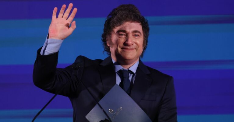 A imagem mostra um homem de terno preto, camisa azul e gravata escura, sorrindo e acenando com a mão direita levantada. Ele segura uma pasta azul com um brasão dourado. O fundo é azul com tons de luz. O homem na imagem foi identificado como Javier Milei, presidente da Argentina