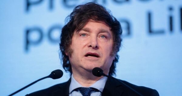 Javier Milei, presidente da Argentina, discursando em um evento. Ele aparece entre dois microfones, vestindo um terno escuro, camisa clara e gravata azul. O fundo exibe um telão com palavras parcialmente visíveis (cripto)