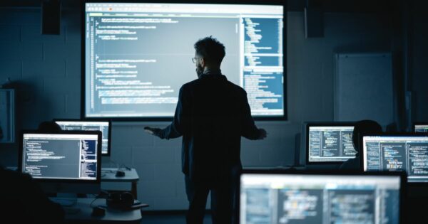 Uma sala escura de aula ou laboratório de informática com múltiplos monitores exibindo código de programação. No centro, um instrutor ou especialista em tecnologia, de costas para a câmera, gesticula enquanto explica algo para os alunos. Uma grande tela de projeção na parede exibe linhas de código, iluminando o ambiente com um brilho azulado. Os alunos, visíveis como silhuetas, estão concentrados em seus computadores. tec