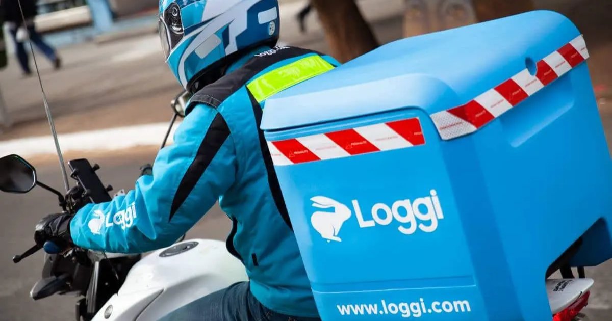 Motoboy da Loggi realizando uma entrega. Ele veste um uniforme azul com a logo da empresa e utiliza um capacete com detalhes brancos. A motocicleta possui um baú azul com o logotipo da Loggi e adesivos refletivos, reforçando a identidade visual da empresa de logística. A imagem transmite a agilidade e eficiência do serviço de entregas urbanas (loggi, motoboy, moto, entrega, logística, plataformas)