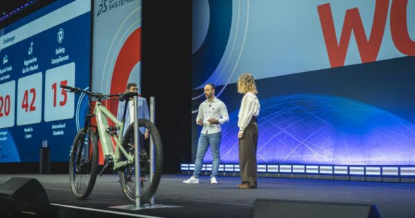 Leandro Timotio da Silva, engenheiro de produtos da Caloi, apresenta-se no palco do 3DExperience World 2025, evento da Dassault Systèmes. Ele está acompanhado de duas outras pessoas, uma mulher e um homem, enquanto fala ao público. No palco, há uma bicicleta exposta em destaque, e ao fundo, um grande telão exibe informações sobre desafios da empresa, incluindo números sobre testes físicos, modelos desenvolvidos e padrões de segurança.