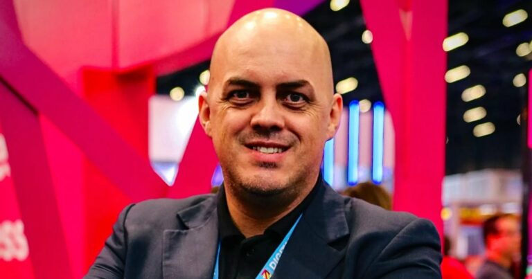 Homem de pele clara, calvo, com barba rala, sorrindo, veste paletó escuro e camiseta preta. Ao fundo, ambiente interno com iluminação artificial e estruturas nas cores rosa e azul, possivelmente em um evento corporativo. A imagem mostra Daniel Sartori, Head de Conectividade Móvel no Brasil da Deutsche Telekom Global Business (Brasil)
