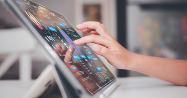 Mão interagindo com um tablet em um suporte, exibindo uma interface digital com gráficos, estatísticas e dados coloridos. A tela apresenta elementos visuais sofisticados, sugerindo uma análise de dados ou painel de controle digital. O fundo está desfocado, destacando o dispositivo e a interação do usuário (desafios)
