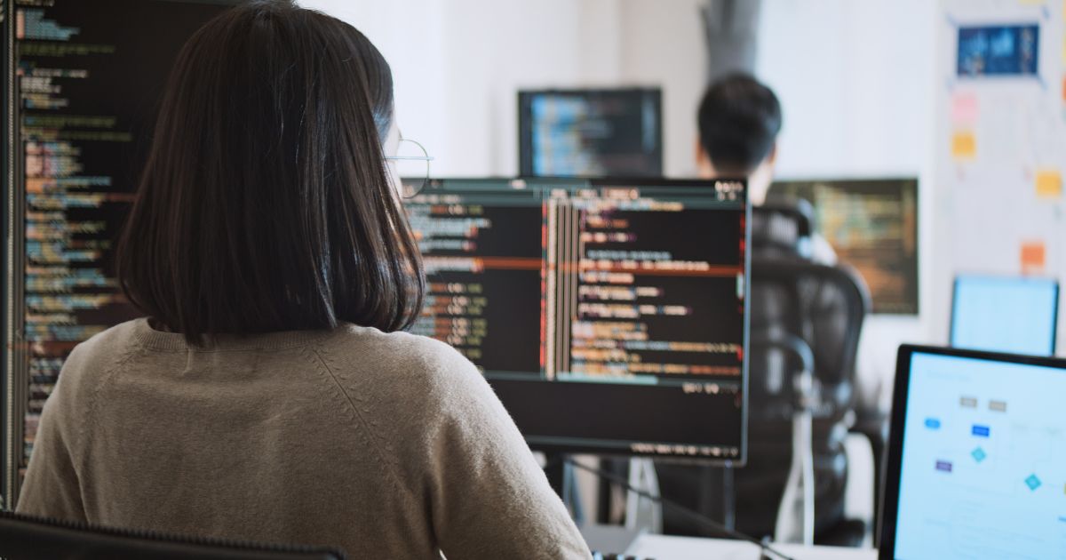 Uma mulher de cabelo escuro e óculos está sentada em frente a vários monitores de computador, trabalhando com código de programação. A tela principal exibe linhas de código coloridas em um ambiente de desenvolvimento. No fundo, outro programador, um homem de cabelo curto, também trabalha em seu computador, e há um quadro branco com anotações e diagramas. O ambiente parece ser um escritório moderno de tecnologia ou uma startup. (contrata, Prompting, dev)
