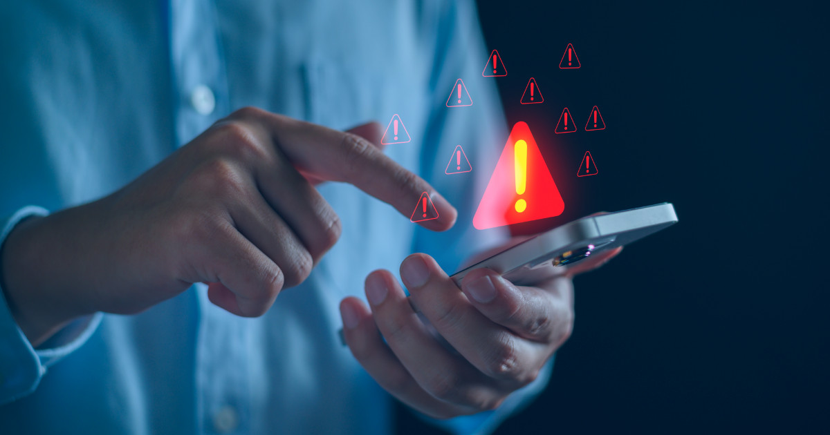 A imagem mostra uma pessoa segurando um smartphone enquanto toca na tela com o outro dedo. Diversos ícones vermelhos de alerta com pontos de exclamação aparecem sobrepostos à cena, simbolizando um aviso ou risco relacionado ao uso do dispositivo. O fundo escuro e desfocado destaca a interação e transmite uma sensação de preocupação ou atenção necessária, sugerindo um possível problema de segurança digital, golpe ou notificação de erro (avisamos, ataques, golpes, Brasil, Gartner)