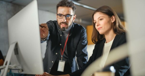 Homem e mulher em ambiente corporativo ou sala de aula moderna, olhando atentos para a tela de um computador. O homem, usando óculos e crachá, aponta para a tela enquanto a mulher observa com expressão concentrada. A cena sugere um momento de orientação ou aprendizado relacionado a tecnologia ou inteligência artificial. operações