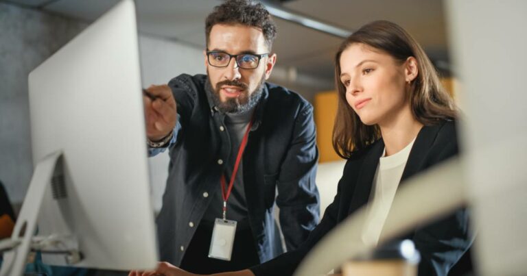 Homem e mulher em ambiente corporativo ou sala de aula moderna, olhando atentos para a tela de um computador. O homem, usando óculos e crachá, aponta para a tela enquanto a mulher observa com expressão concentrada. A cena sugere um momento de orientação ou aprendizado relacionado a tecnologia ou inteligência artificial. operações