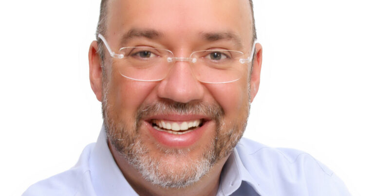 Jorge Luís Cordenonsi, CIO da Compass Latin America e GRSA, sorrindo, usando óculos de armação transparente, camisa azul clara e fundo branco.