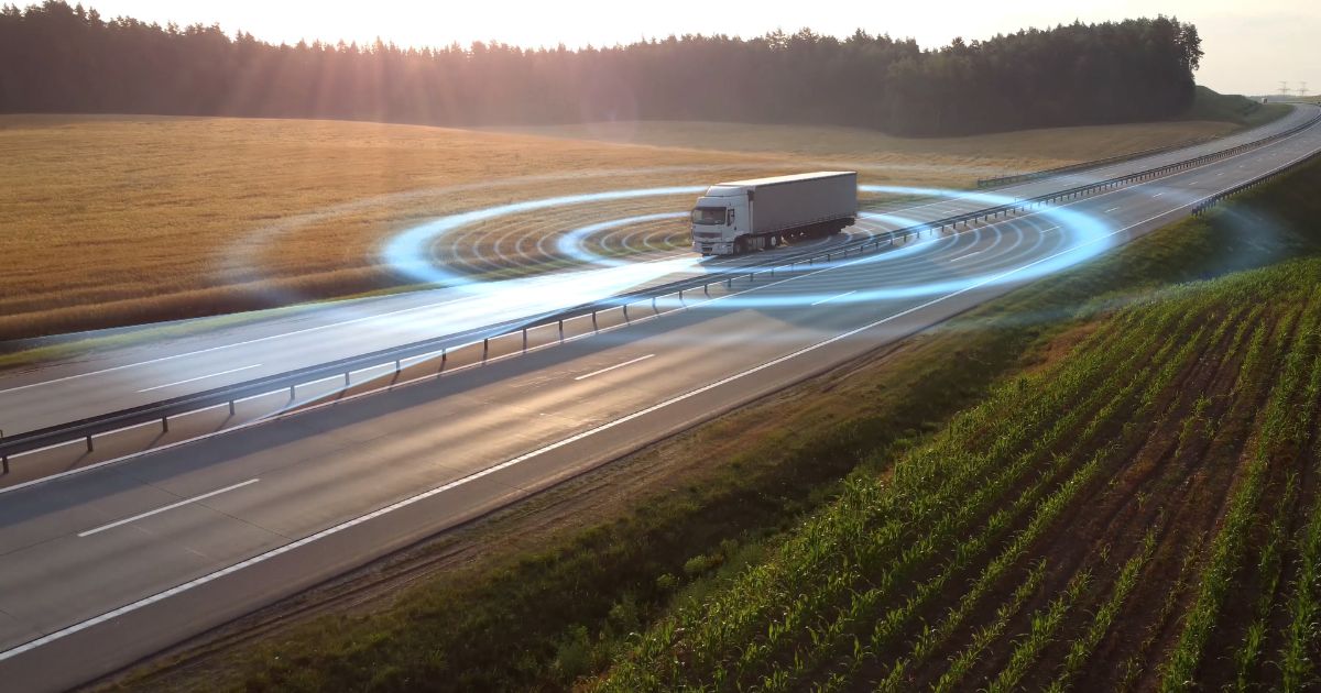 Um caminhão branco trafega em uma rodovia cercada por campos verdes e dourados ao pôr do sol. Há um efeito gráfico azul simulando ondas ao redor do veículo, sugerindo tecnologia de sensores ou conectividade digital na estrada (cimento, reduzir)