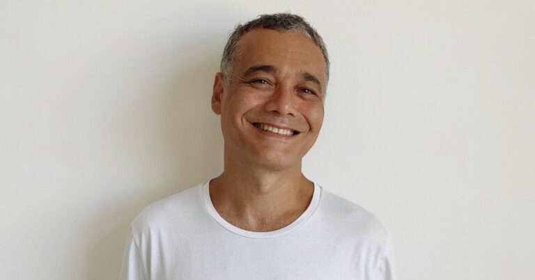 Sidarta Ribeiro sorri levemente em frente a uma parede clara. Ele veste uma camiseta branca e tem cabelo curto e grisalho. Foto: Luiza Mugnol Ugarte (tecnologia)