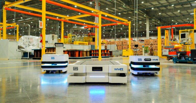 Imagem de uma fábrica moderna com três robôs móveis autônomos (AMRs) da WEG em parceria com a Senior, posicionados em destaque no centro do ambiente industrial. Os robôs, identificados pelas marcas “WEG” e “WMR”, estão em um chão limpo e bem iluminado, cercados por estruturas metálicas amarelas e equipamentos industriais ao fundo, ilustrando uma solução de automação voltada para logística e movimentação de materiais.