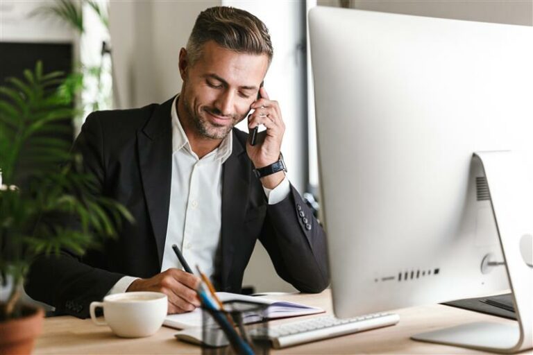 Homem de terno sentado à mesa, falando ao telefone e escrevendo em um caderno, com um monitor de computador iPlace Corporativo, uma xícara de café e uma planta por perto