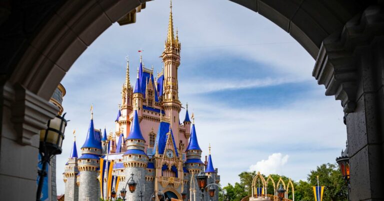 Castelo da Cinderela no Magic Kingdom, no Walt Disney World, Orlando, nos Estados Unidos, enquadrado por um arco de pedra em primeiro plano. O castelo tem torres azul-royal e detalhes dourados, sob um céu azul com algumas nuvens.